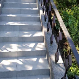 Zewnętrzne schody wyłożone szarymi płytkami, z metalową balustradą o geometrycznym wzorze, oświetlone słonecznym światłem, z widoczną zielenią w tle.