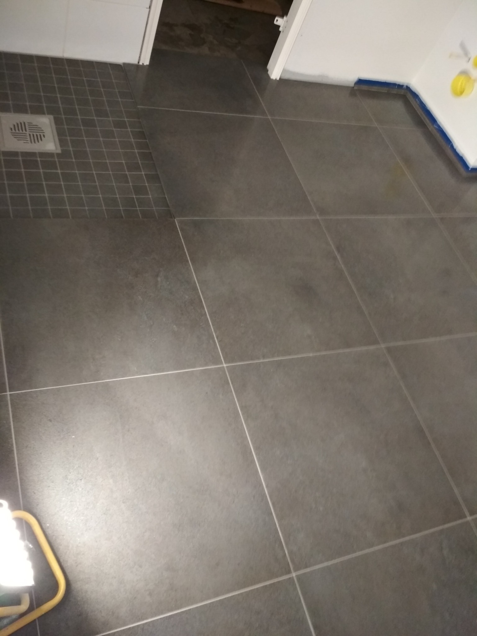 Ułożone szare płytki podłogowe w łazience we Wrocławiu, widoczny odpływ liniowy i zabezpieczona folią ściana przed malowaniem.