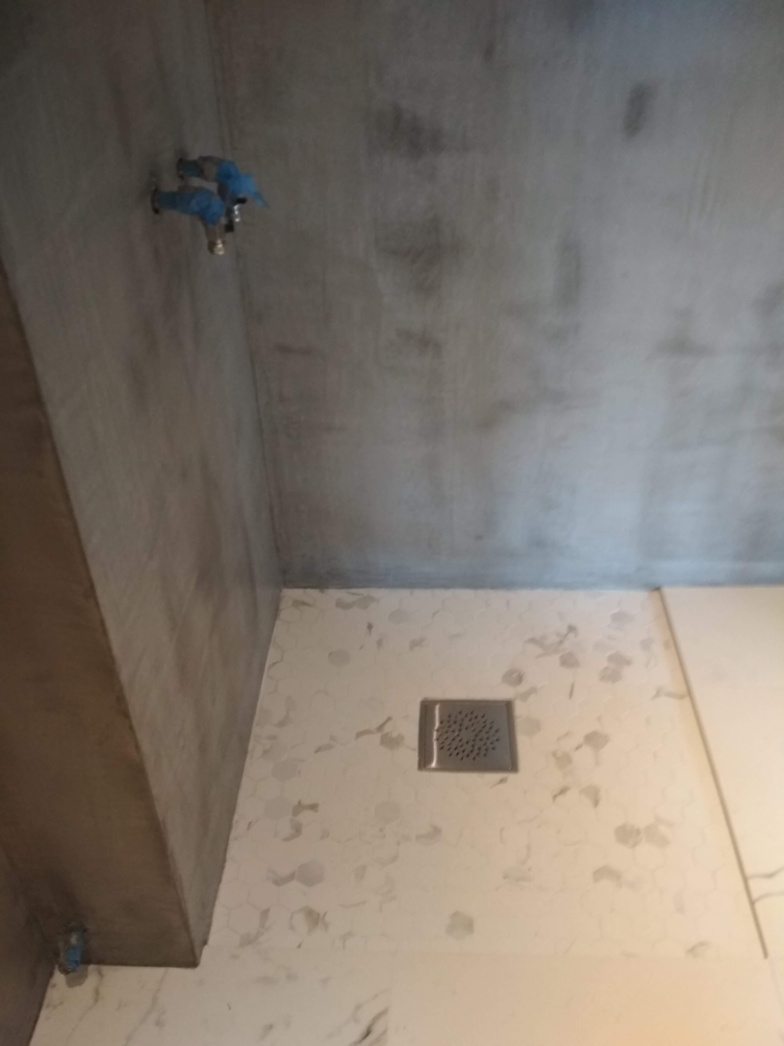 Surowy beton na ścianie nad fragmentem łazienki z ułożonymi płytkami heksagonalnymi w odcieniach bieli i szarości, z odpływem liniowym w podłodze i zabezpieczonymi niebieską folią przyłączami wodnymi.