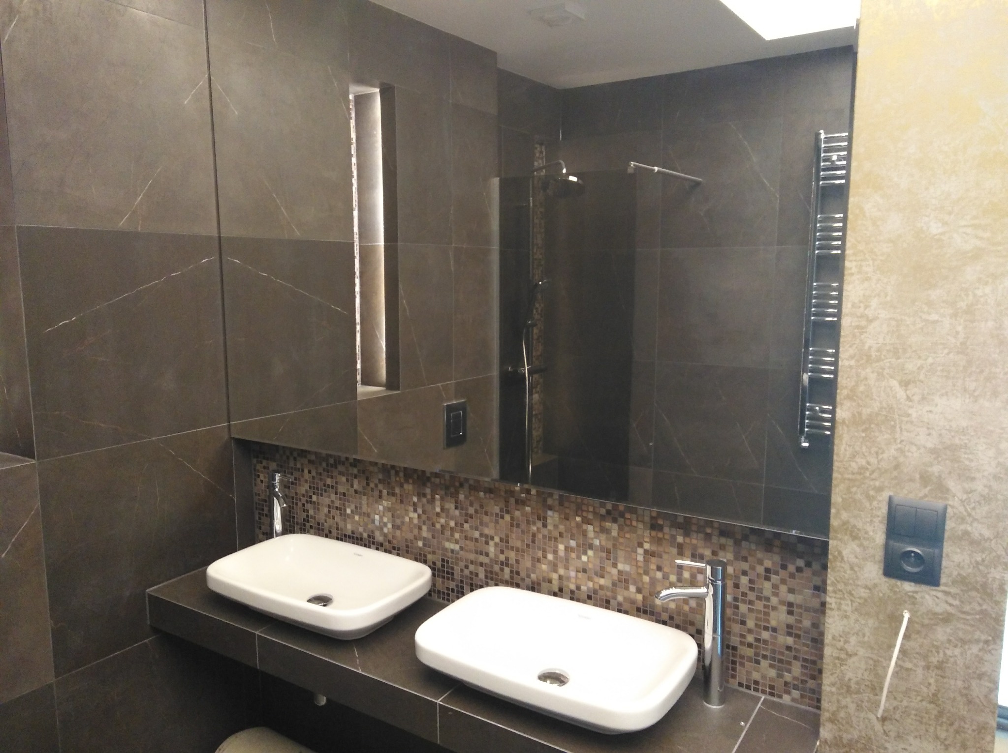 Nowoczesna łazienka z dwoma białymi umywalkami nablatowymi, ciemnymi płytkami imitującymi marmur na ścianach, mozaiką nad umywalkami i dużym lustrem.