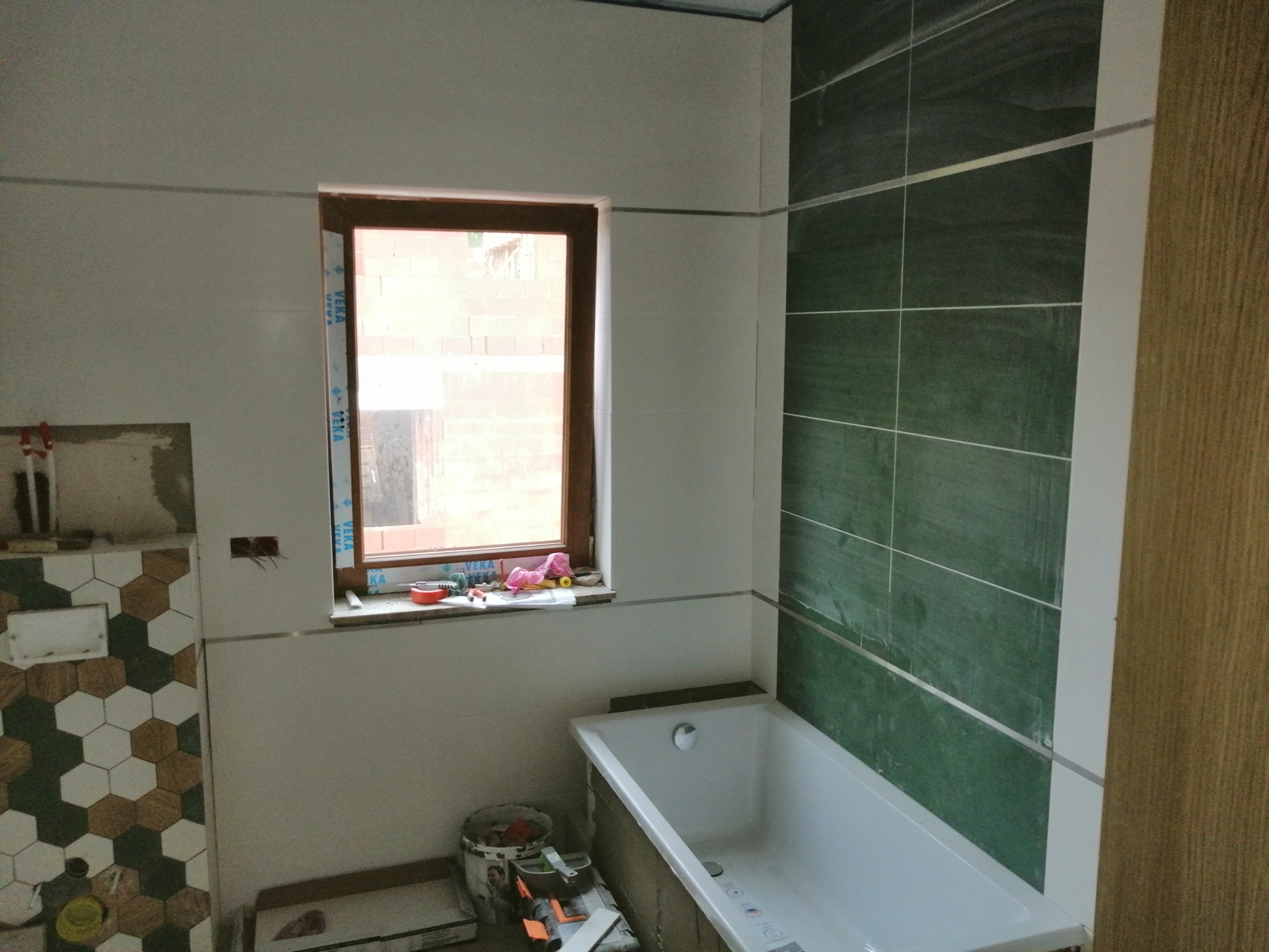 Wnętrze łazienki w trakcie prac wykończeniowych: zielone płytki na ścianie obok wanny, fragment ściany z heksagonalnymi płytkami, okno z zabawkami na parapecie, widoczne narzędzia i materiały...