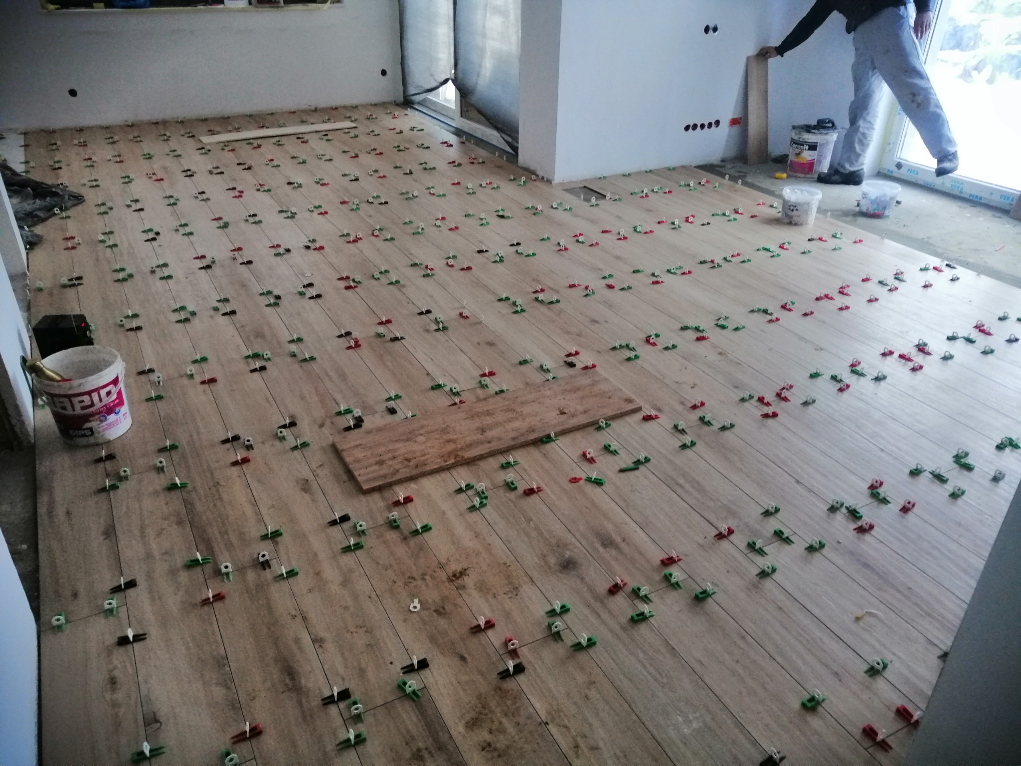 Układanie podłogi z płytek imitujących drewno, z użyciem klipsów poziomujących w kolorach czerwonym, zielonym i czarnym, widoczne wiaderka z klejem i osoba w tle.