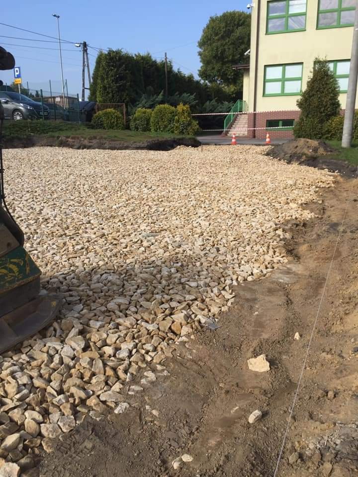 Przygotowanie podłoża z kruszywa na posesji, widoczny ubijak wibracyjny i wytyczone granice przyszłej powierzchni. Budynek w tle.