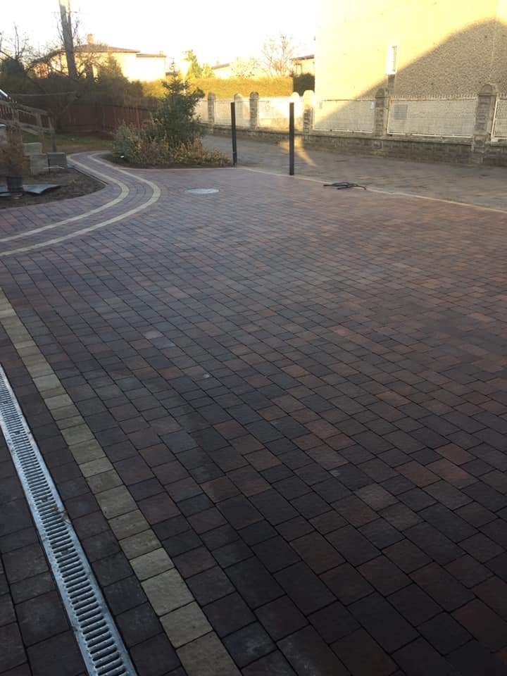 Ułożona, brązowo-szara kostka brukowa z liniowym odwodnieniem po lewej stronie, tworząca ścieżkę wokół rabaty z krzewami, z budynkiem w tle.