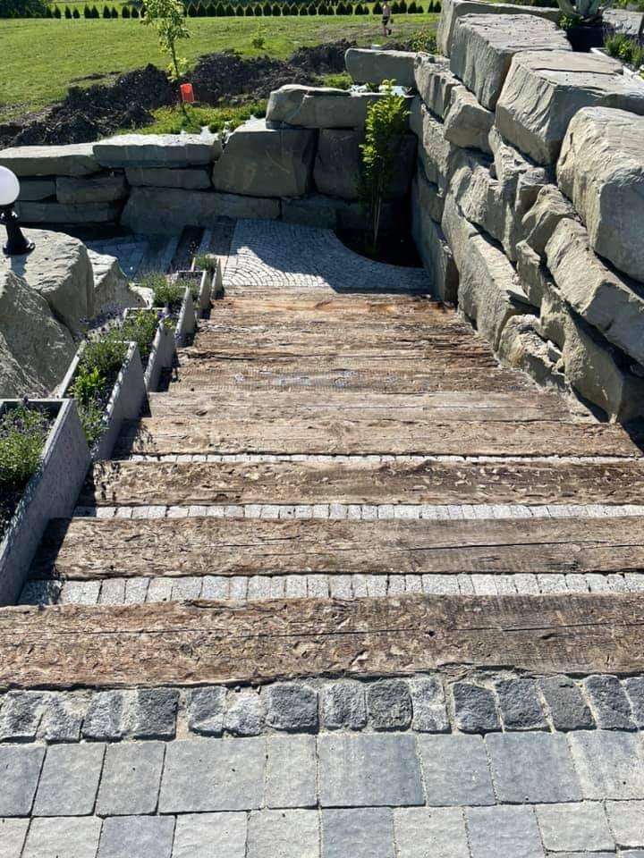 Schody zewnętrzne z drewnianych bali i kostki granitowej, z donicami po bokach, otoczone kamiennym murem oporowym i trawnikiem.