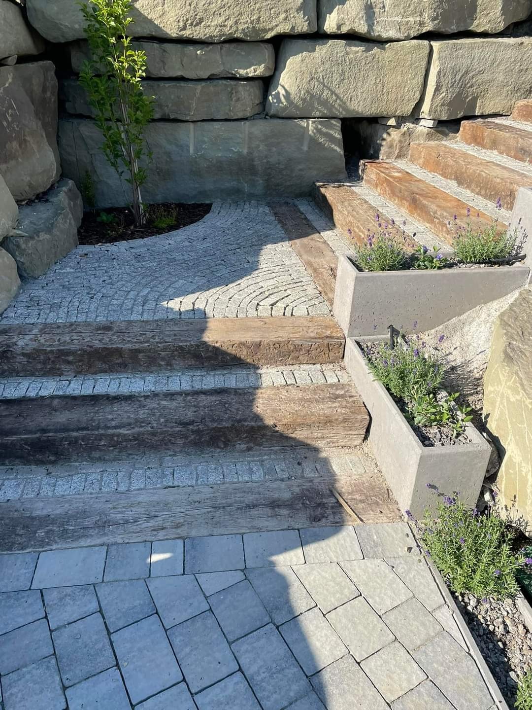 Kamienne schody z drewnianymi stopniami i brukowaną ścieżką, otoczone roślinnością i kamiennym murem oporowym.