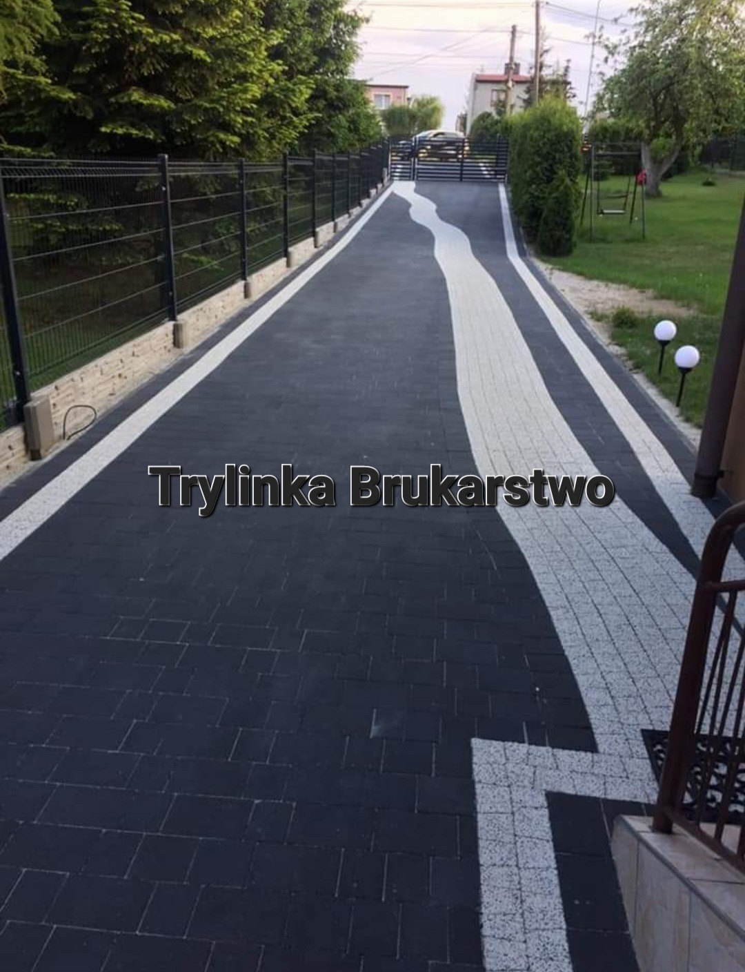 Czarna kostka brukowa ułożona wzdłuż podjazdu z białym pasem pośrodku i białymi obrzeżami, widok z góry na posesję z ogrodzeniem i zielenią.