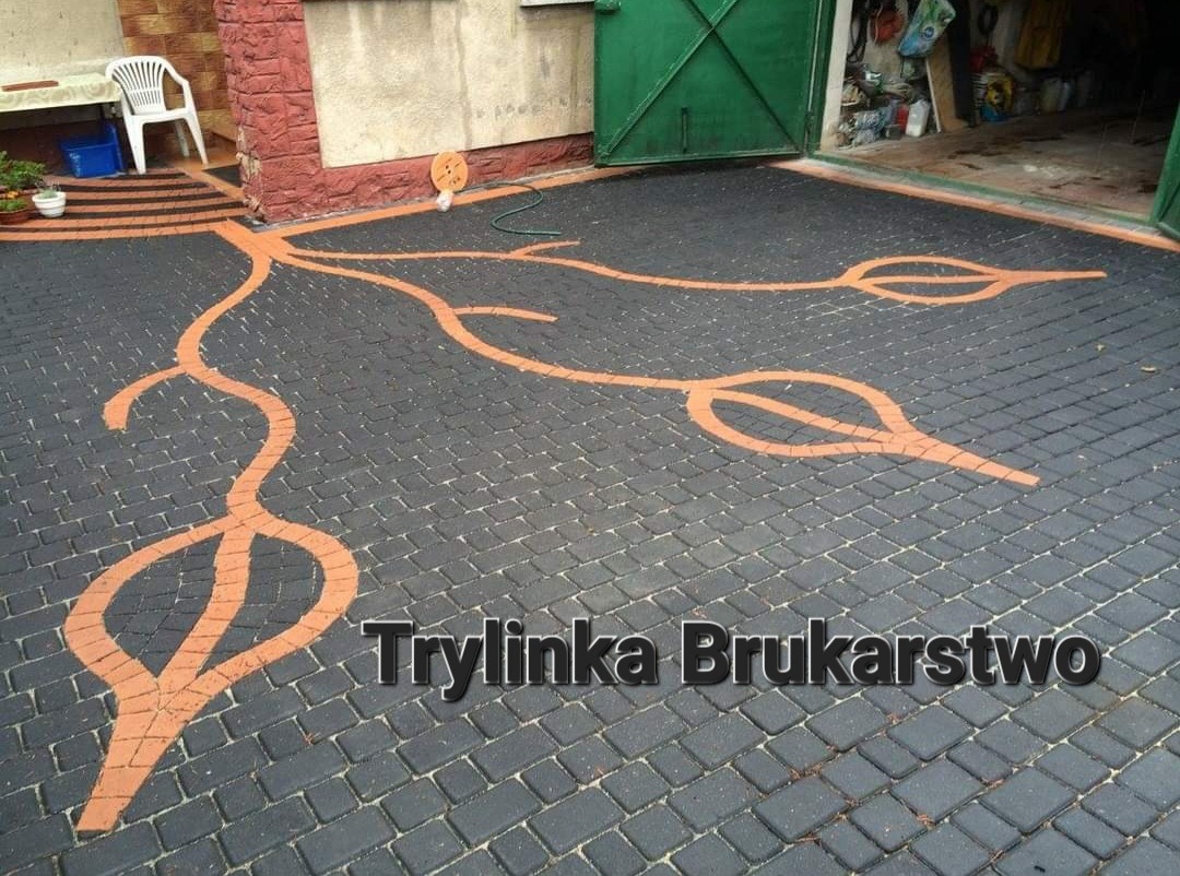 Czarna kostka brukowa ułożona w oryginalny wzór z pomarańczowymi akcentami przypominającymi korzenie drzewa i kształty liści, prowadząca do wejścia do domu z zielonymi drzwiami garażowymi.