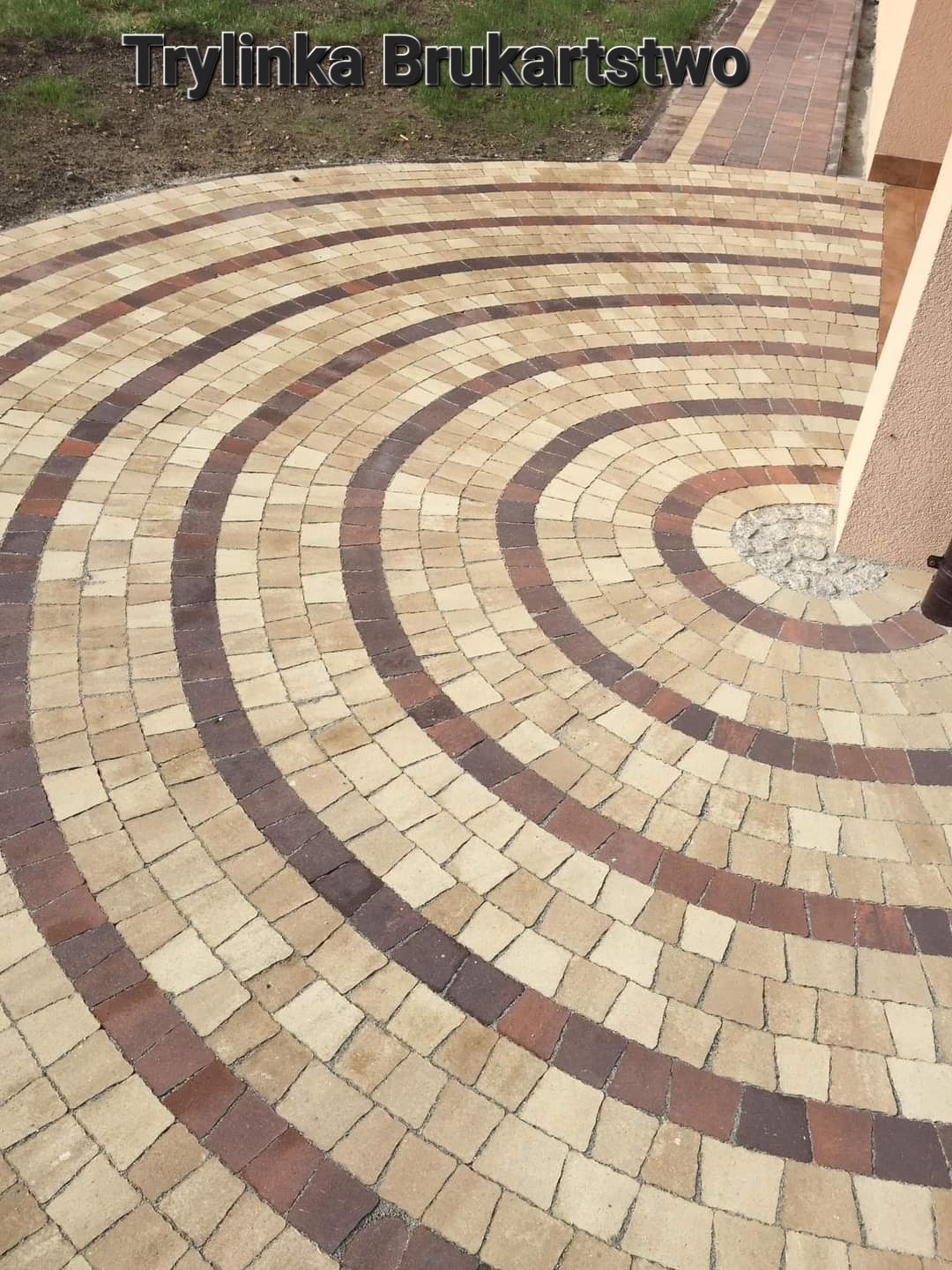Okrągły wzór z kostki brukowej w odcieniach beżu i brązu, ułożony koncentrycznie wokół centralnego punktu, z widocznym fragmentem budynku w tle.