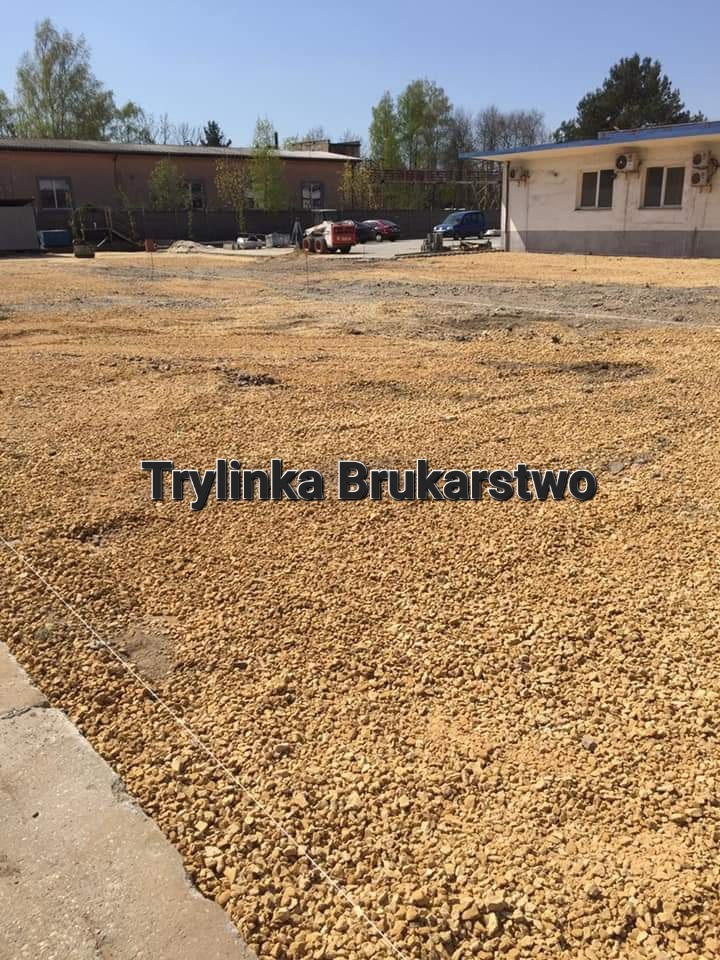 Przygotowany teren pod układanie kostki brukowej, podsypka z kruszywa ograniczonego krawężnikiem, widoczna wytyczona niwelacja terenu sznurkiem, w tle budynki i maszyny.