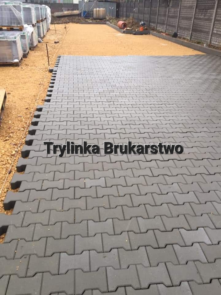 Układanie szarej kostki brukowej typu 'H' na podsypce piaskowej, widoczne palety z kostką w tle i przygotowany teren.