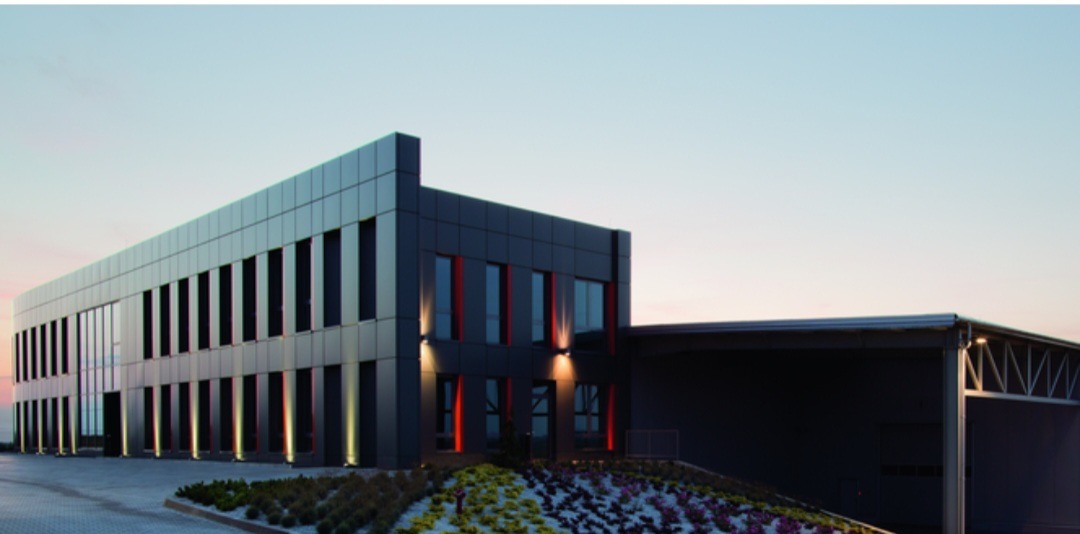 Nowoczesny budynek biurowy z elewacją w odcieniach szarości, oświetlony lampami zewnętrznymi, z widocznym fragmentem parkingu i rabatą z roślinami na pierwszym planie, sfotografowany o zmierzchu.