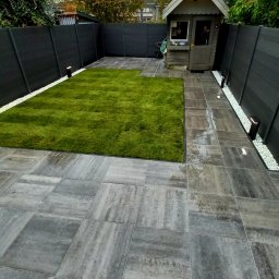 Q-74 Tuinen - Nowoczesny ogród z równo przyciętą trawą, wyłożony szarymi płytami chodnikowymi, z ciemnym ogrodzeniem i małym domkiem narzędziowym w tle.