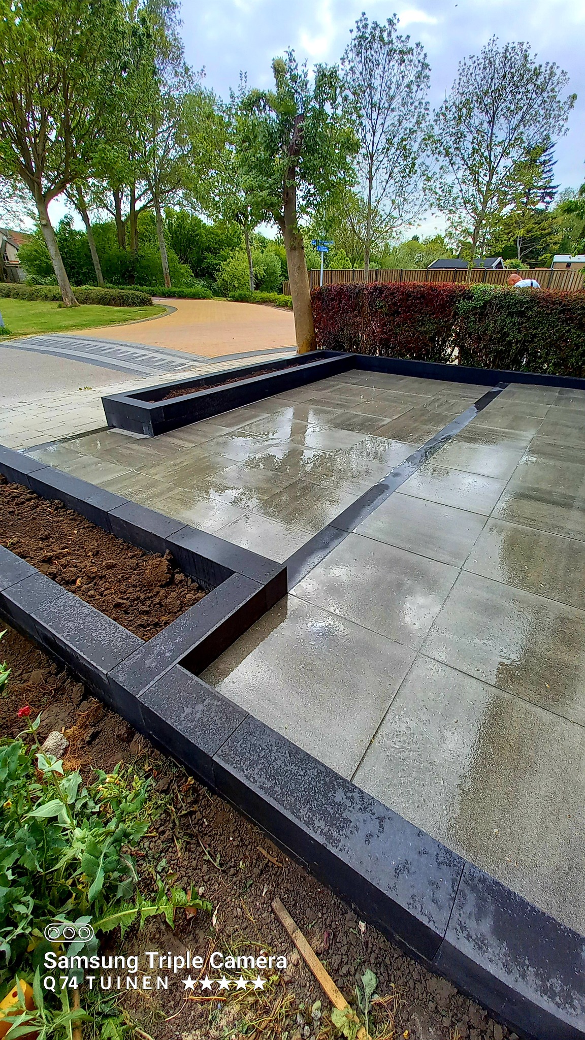 Ułożona, mokra nawierzchnia z szarych płyt obramowana czarnymi krawężnikami, tworząca rabaty z ziemią i przestrzeń do nasadzeń, w tle widoczne drzewa i krzewy.