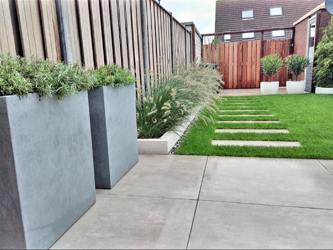 Nowoczesny ogród z betonową ścieżką, wysokimi donicami z ziołami i dekoracyjnymi trawami, ogrodzony drewnianym płotem.