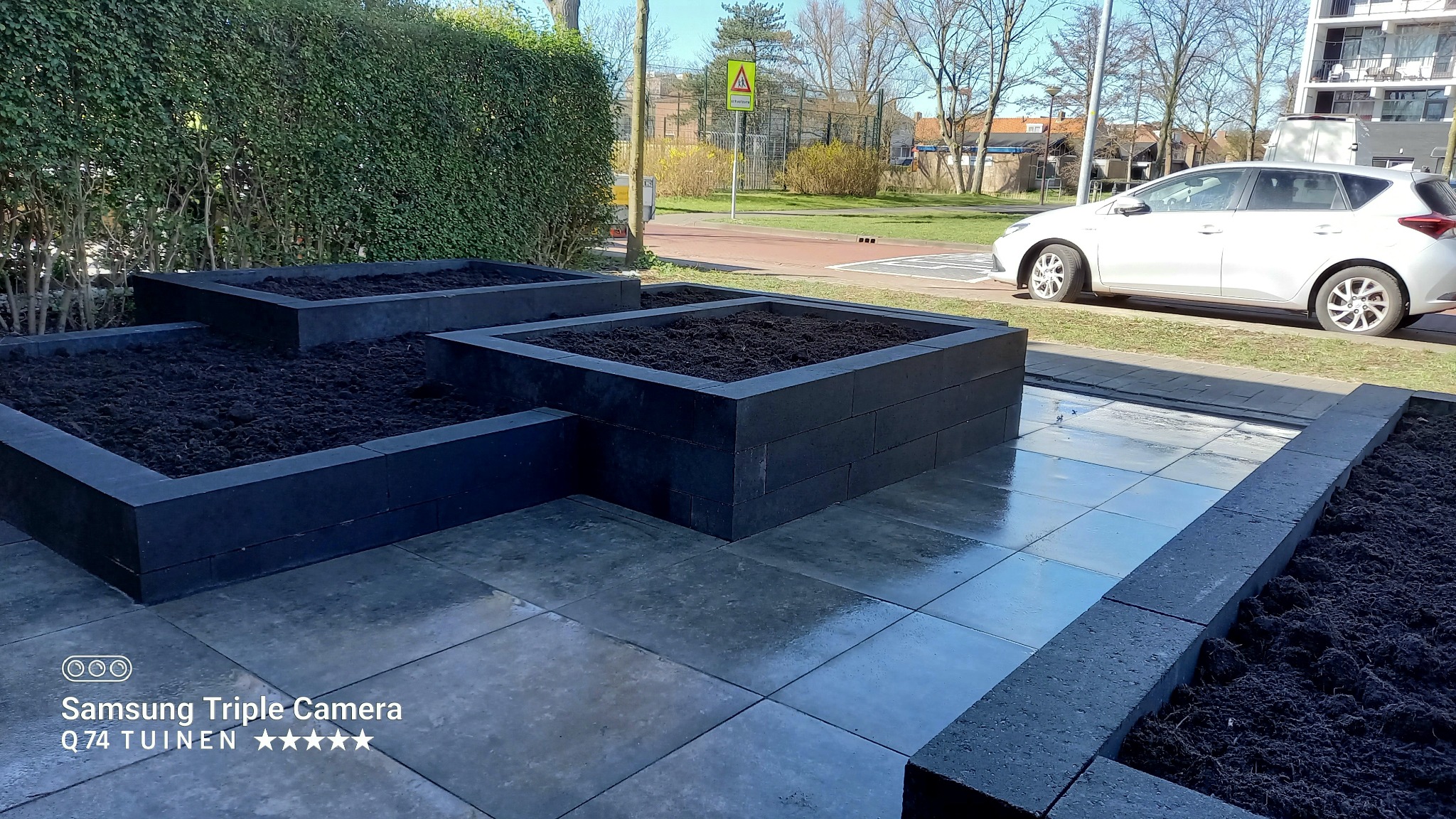Podwyższone rabaty z ciemnych betonowych bloczków, wypełnione ziemią, ułożone na szarym bruku; w tle żywopłot, ulica i zaparkowany samochód.
