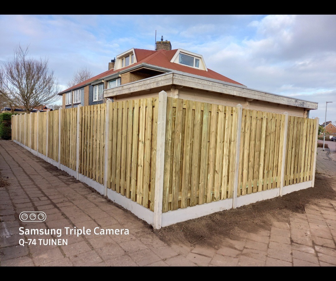 Nowo postawione ogrodzenie panelowe z jasnego drewna z betonowymi słupkami i podmurówką, widoczne z poziomu chodnika przed domem z czerwonym dachem w pochmurny dzień.