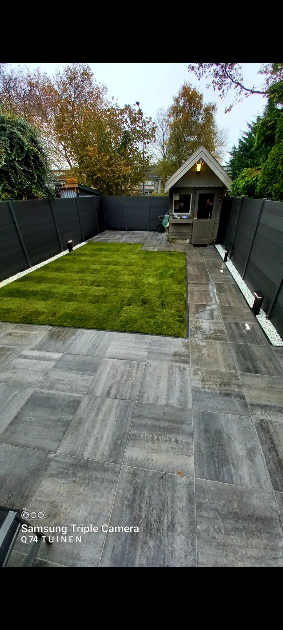 Nowoczesny ogród z równo przyciętą trawą, wyłożony szarymi płytami chodnikowymi, z ciemnym ogrodzeniem i małym domkiem narzędziowym w tle.