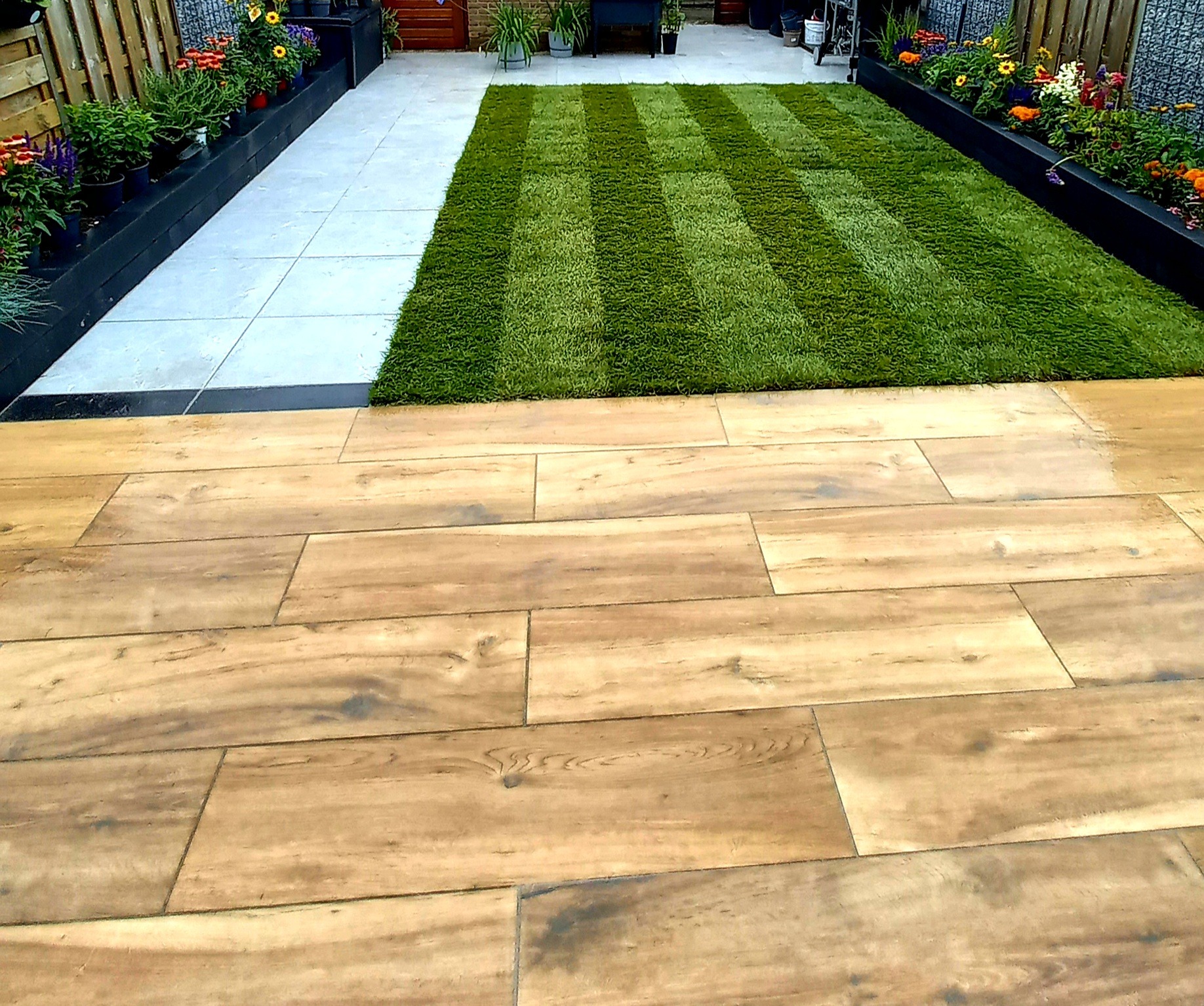 Ułożona trawa w paski na tle jasnych płyt chodnikowych i drewnopodobnej terakoty, rabaty kwiatowe wzdłuż ogrodzenia.