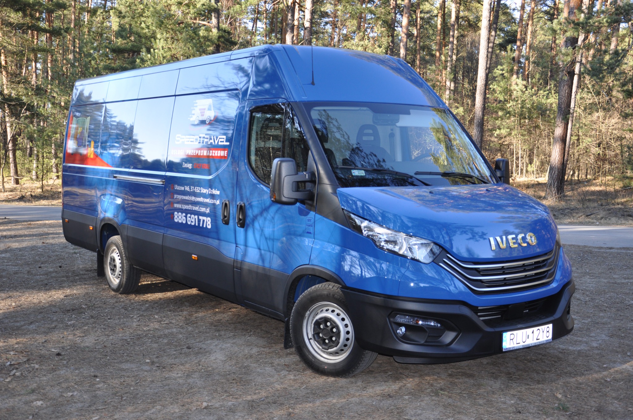 Niebieski bus marki Iveco z logo firmy przeprowadzkowej Speed Travel, zaparkowany na leśnym parkingu w słoneczny dzień.