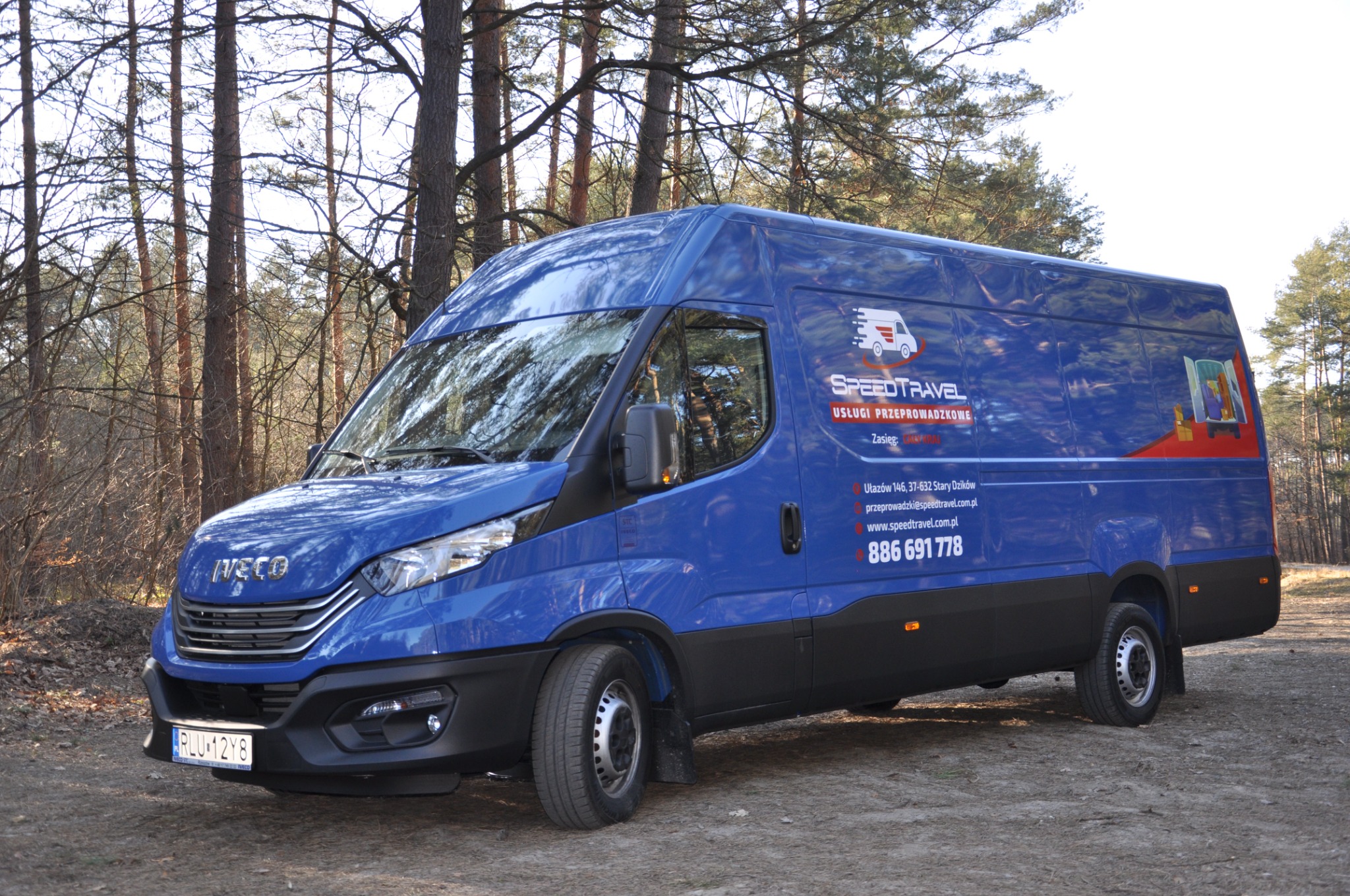 Niebieski samochód dostawczy Iveco z logo firmy SPEEDTRAVEL na tle lasu, z widoczną tablicą rejestracyjną RLU 12Y8.