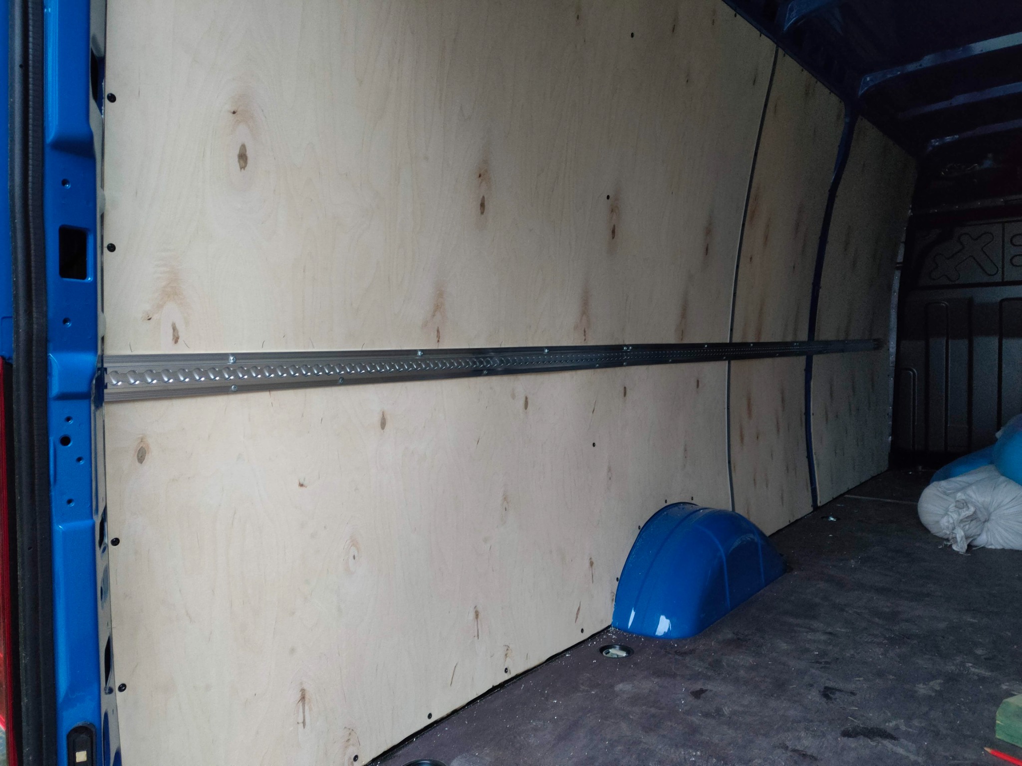 Wnętrze częściowo wykończonego busa z drewnianą zabudową ścian i szyną mocującą ładunek, widoczny niebieski nadkole i worki z materiałem.