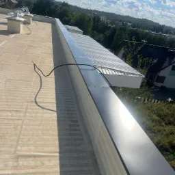 Jasny, płaski dach z blachy trapezowej i systemem odprowadzania wody, widok z góry, na tle zabudowań i błękitnego nieba z chmurami.