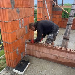 WorkBud - Murarz w trakcie wznoszenia ściany z czerwonej cegły ceramicznej. Widoczne narzędzia i akcesoria budowlane, drabina oraz fragmenty konstrukcji. Praca w toku, etap budowy.