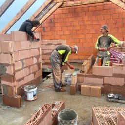WorkBud - Murowanie ścian domu z czerwonej cegły ceramicznej. Dwóch murarzy pracuje, trzeci stoi obok. Widoczne narzędzia i wiaderka z zaprawą. Konstrukcja dachu w tle.