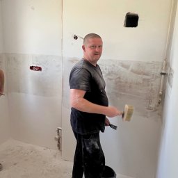 WorkBud - Mężczyzna z pędzlem w trakcie nakładania gładzi na ścianę w remontowanym pomieszczeniu. Widoczne ślady po instalacjach i przygotowania do malowania.