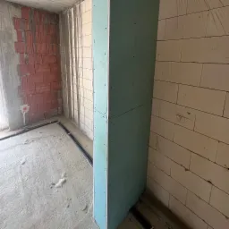 Wnętrze w trakcie prac wykończeniowych: fragment ściany z cegły, pionowa konstrukcja z płyty karton-gips, instalacja elektryczna przy podłodze i surowy beton.