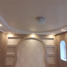 Jasne wnętrze z dekoracyjnym sufitem podwieszanym, oświetleniem punktowym i wnękami ściennymi w kształcie łuków, ściany pokryte tapetą w delikatny wzór.