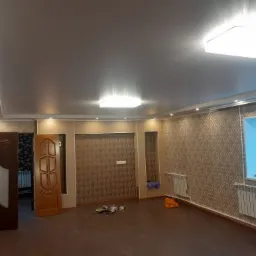 Pokój po remoncie z brązową podłogą, wzorzystą tapetą, białymi grzejnikami, dwoma niedopasowanymi drzwiami i oświetleniem sufitowym.