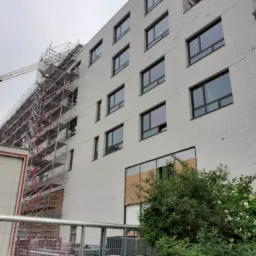 Wielopiętrowy budynek w trakcie budowy z widocznymi rusztowaniami i niezabezpieczonymi oknami, częściowo zasłonięty zielenią.