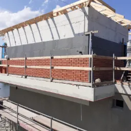 Budowa domu nad wodą: wznoszenie ścian z cegły klinkierowej na betonowej konstrukcji, widoczne rusztowania i elementy konstrukcji dachu.