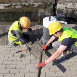Dwóch pracowników w kaskach i kamizelkach odblaskowych naprawia metalowy słupek przy nabrzeżu, widoczny mini-ładowarka i ceglany mur z porostami w tle.