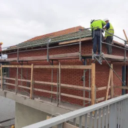 Budowa domu z czerwonej cegły na platformie nad wodą, robotnicy na rusztowaniu układają dachówkę.
