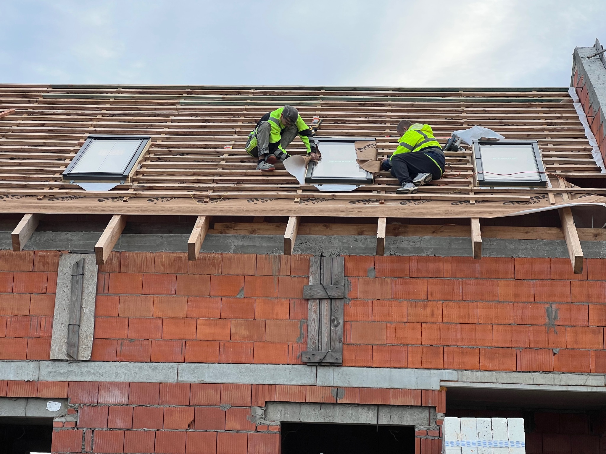Montaż okien dachowych FAKRO na drewnianej konstrukcji dachu, widok z dołu na budynek w trakcie budowy z czerwonej cegły, dwóch dekarzy w odblaskowych kamizelkach.