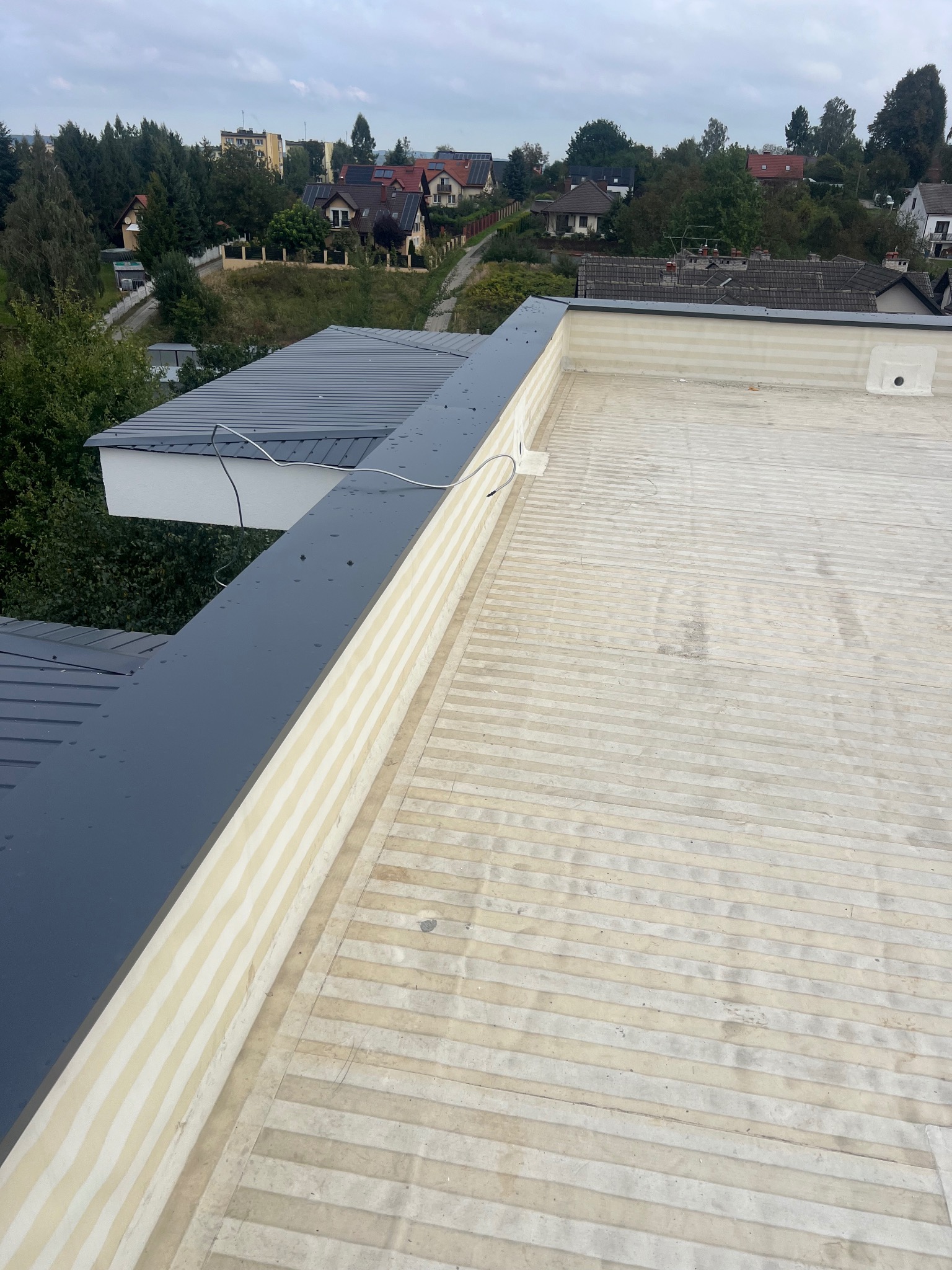 Wykończony płaski dach z szarą obróbką blacharską i widokiem na zabudowę jednorodzinną w tle. Detal konstrukcyjny, poziome ułożenie elementów.