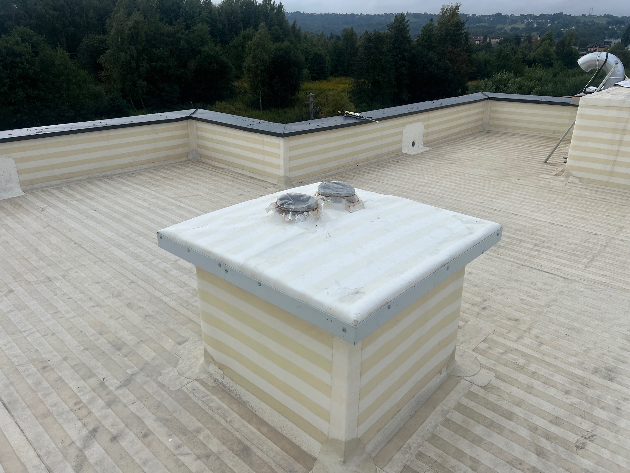 Biało-beżowy płaski dach z wykończoną obudową komina i obróbkami blacharskimi, z widocznymi detalami izolacji i pokrycia. W tle zieleń drzew.