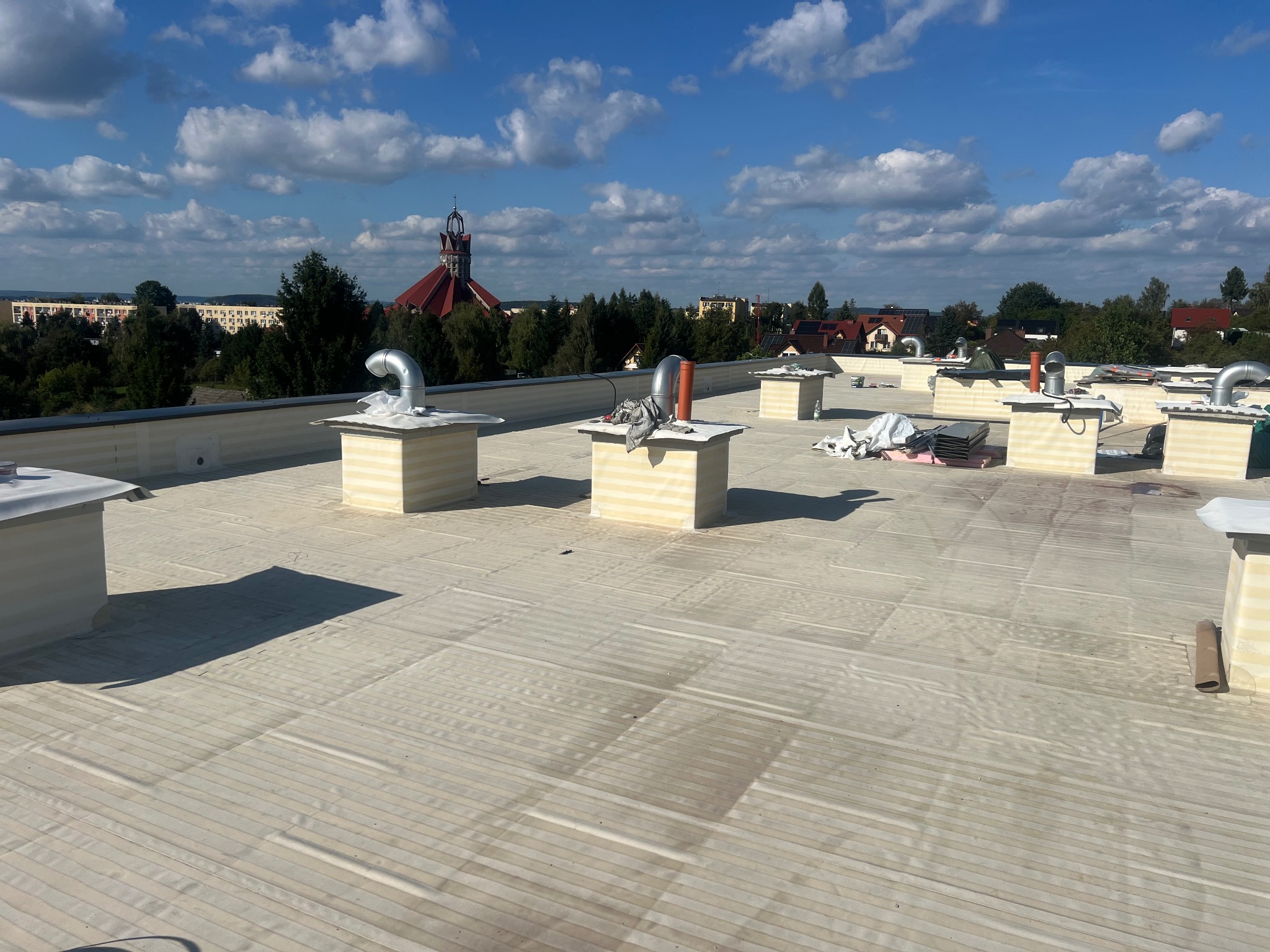 Wykończony dach płaski z odpowietrznikami i kominkami wentylacyjnymi na tle błękitnego nieba i zabudowań miejskich w oddali. Widoczne detale pokrycia dachowego.