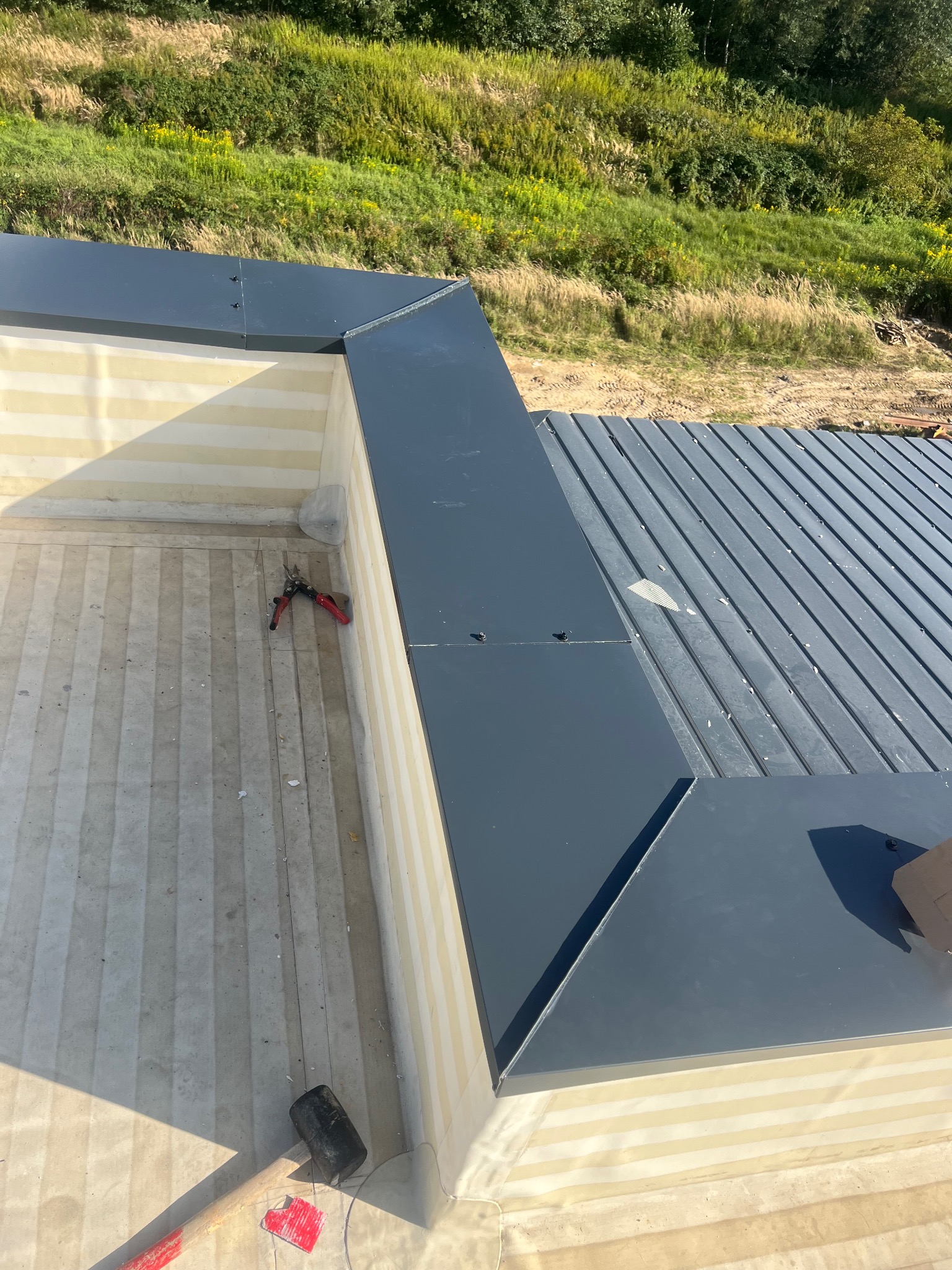 Montaż blachy trapezowej na dachu budynku. Widoczne narzędzia: nożyce do blachy i młotek. W tle zieleń. Dach pokryty blachą w kolorze grafitowym.