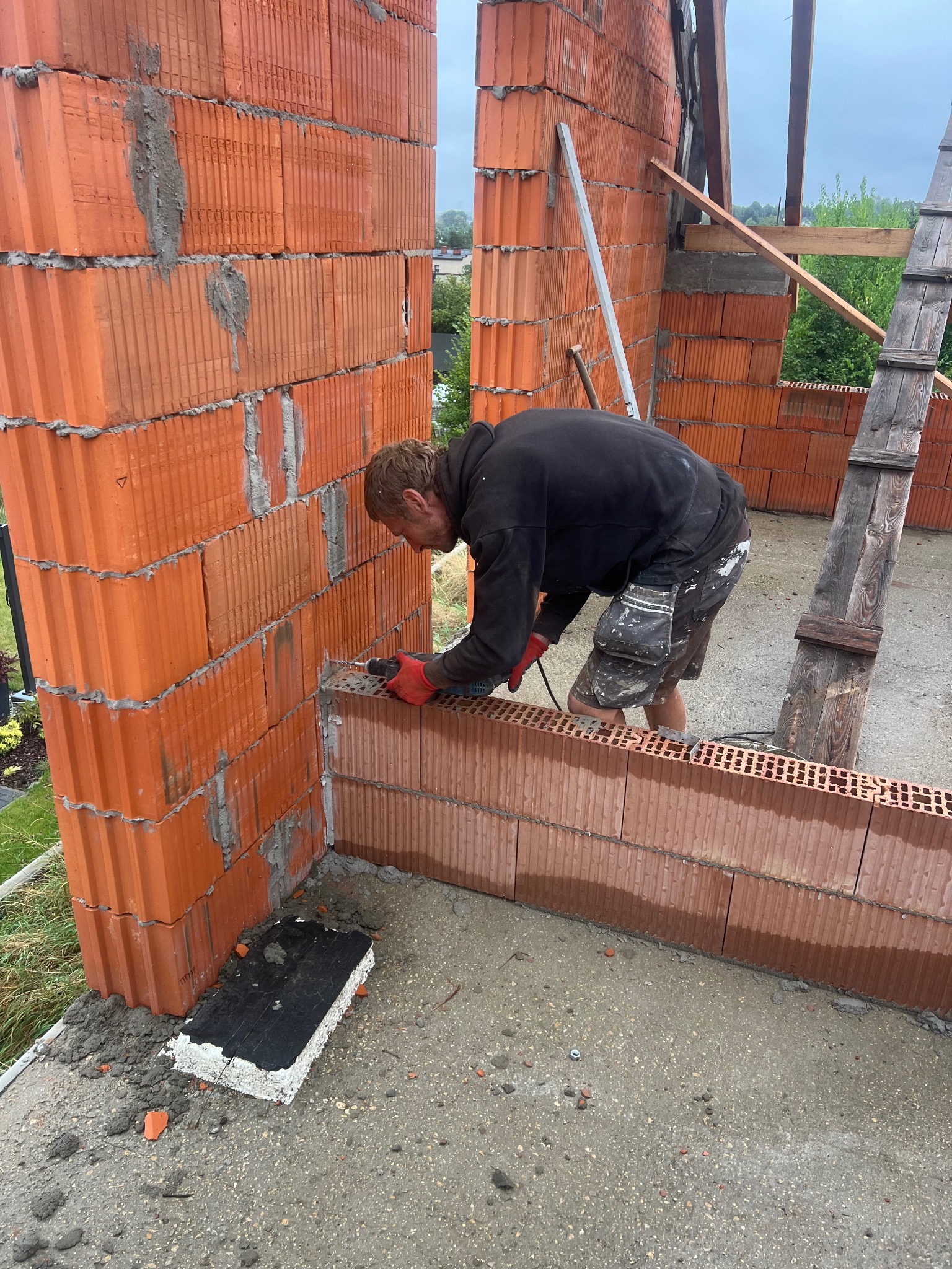 Murarz w trakcie wznoszenia ściany z czerwonej cegły ceramicznej. Widoczne narzędzia i akcesoria budowlane, drabina oraz fragmenty konstrukcji. Praca w toku, etap budowy.