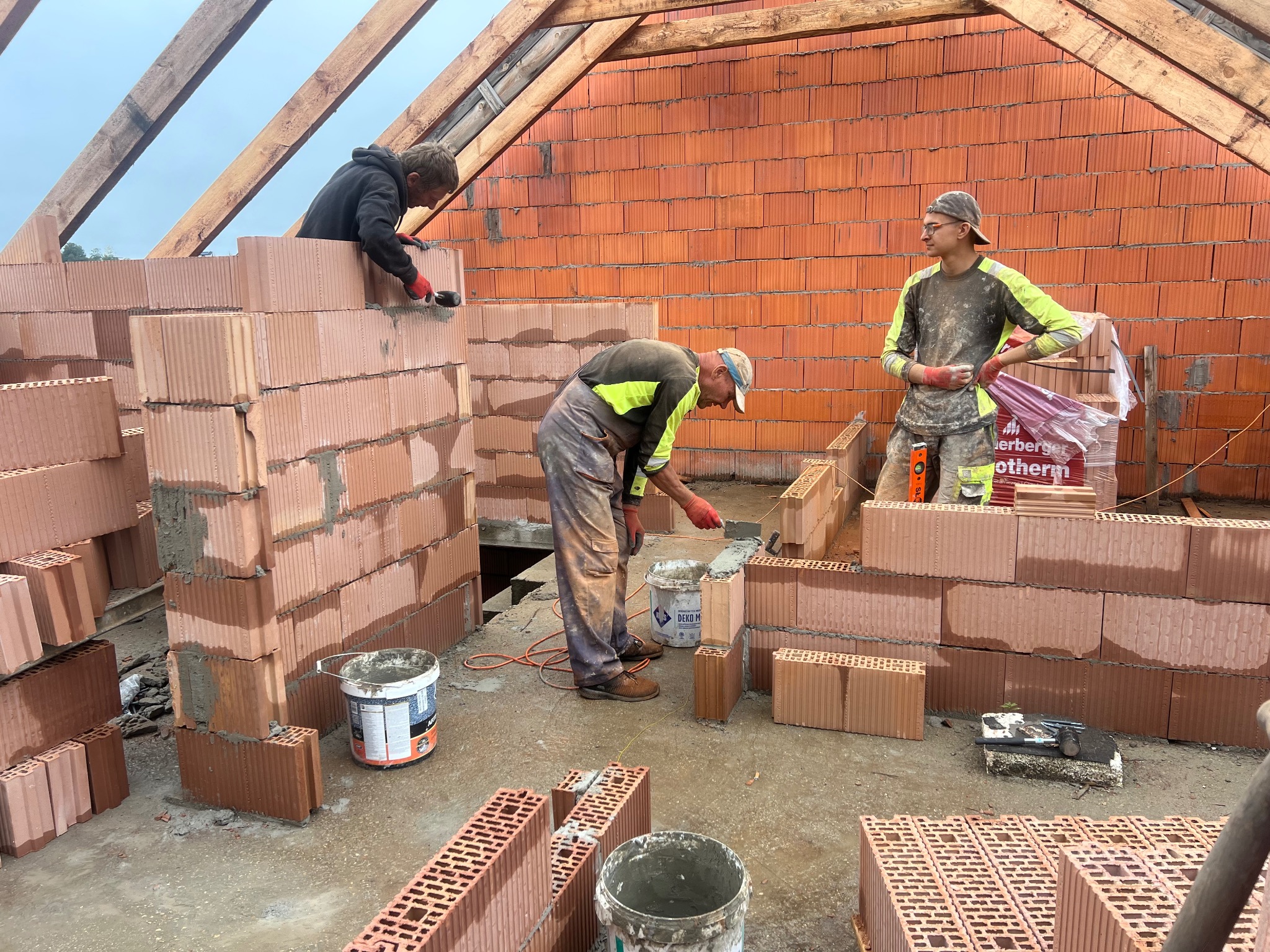 Murowanie ścian domu z czerwonej cegły ceramicznej. Dwóch murarzy pracuje, trzeci stoi obok. Widoczne narzędzia i wiaderka z zaprawą. Konstrukcja dachu w tle.