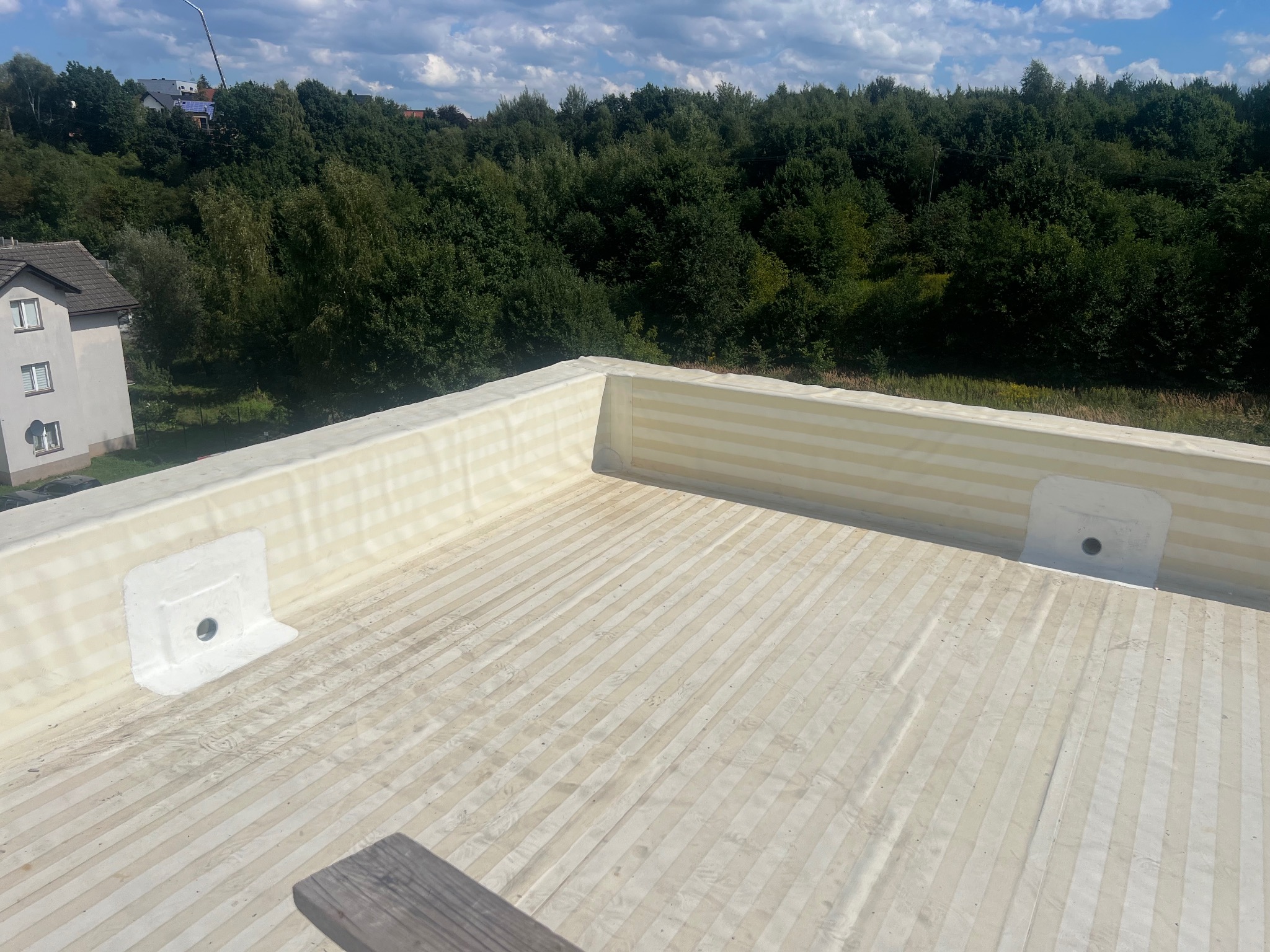 Wykończony płaski dach z izolacją termiczną i odpływami, widok z góry na tle zieleni i błękitnego nieba z chmurami. Detal izolacji i odpływów.