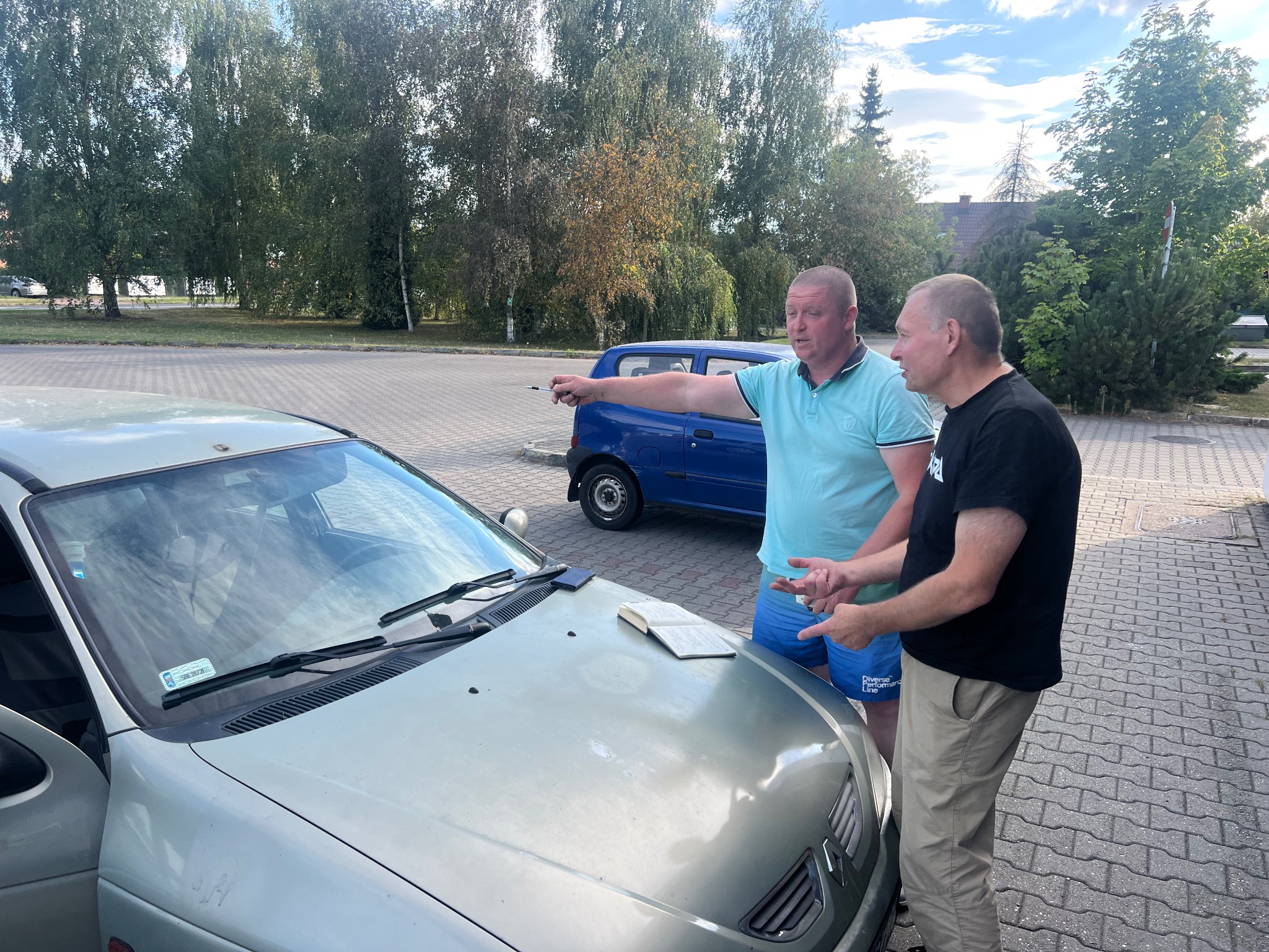 Dwóch mężczyzn w trakcie rozmowy przy samochodzie w Krakowie. Jeden z nich wskazuje palcem w dal, drugi gestykuluje. Na masce leży otwarta książka.