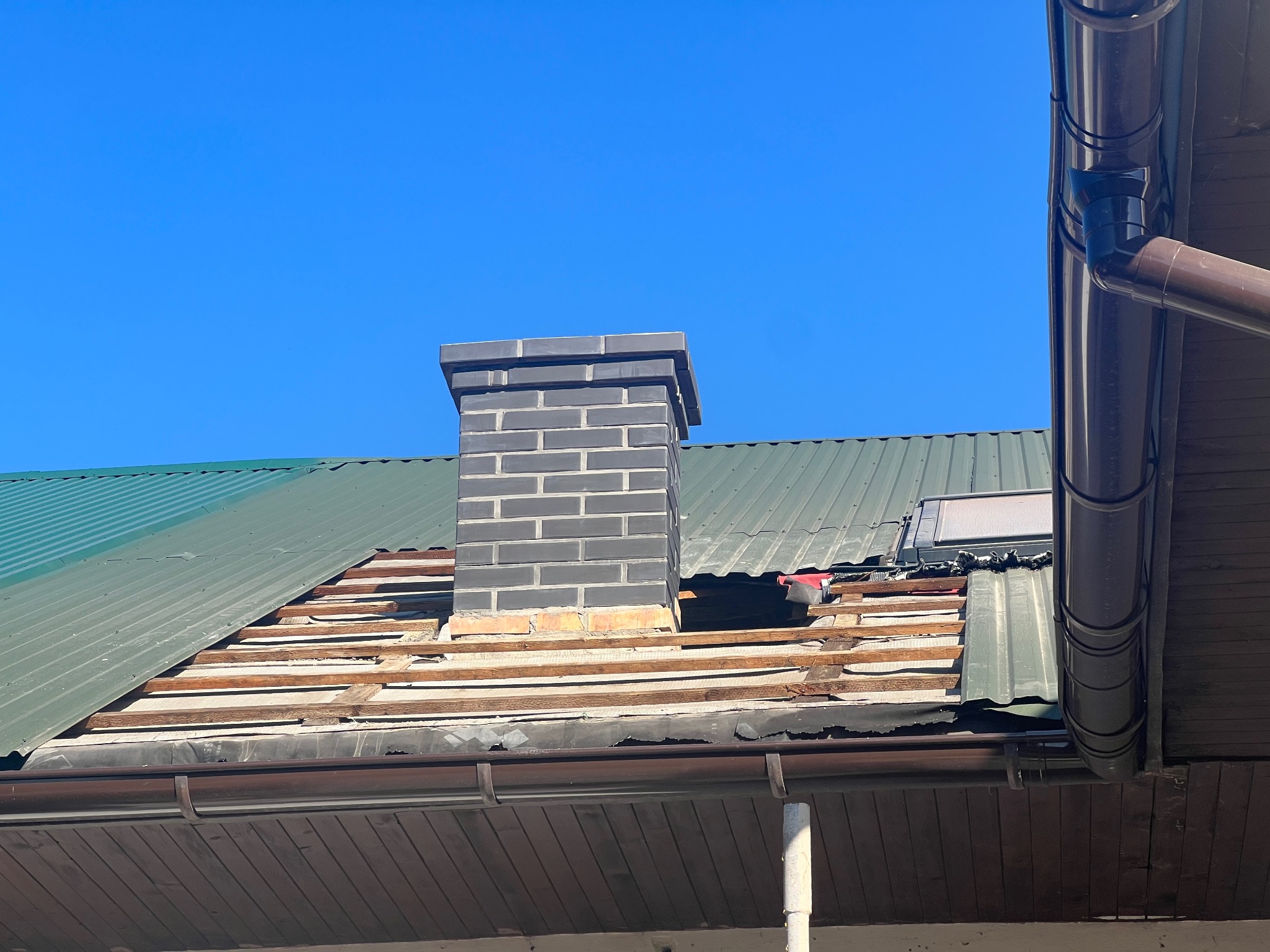 Uszkodzony dach z widocznymi deskami, częściowo pokryty zieloną blachą. Komina z szarej cegły. Brązowa rynna. Błękitne niebo w tle. Widok z dołu.