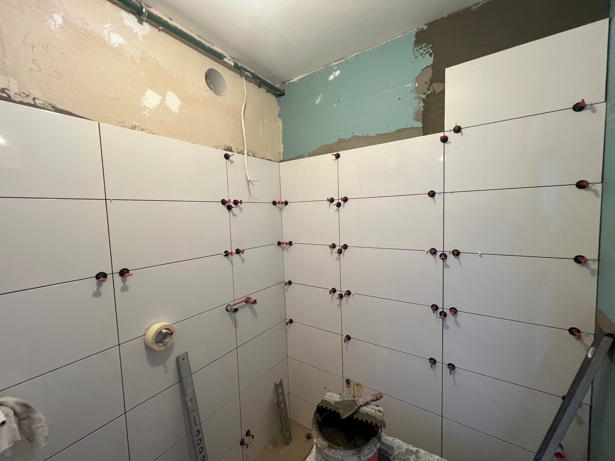 Układanie białych płytek w łazience, widoczne systemy poziomowania. Częściowo wykończone ściany, przygotowane podłoże. Użyte narzędzia glazurnicze.