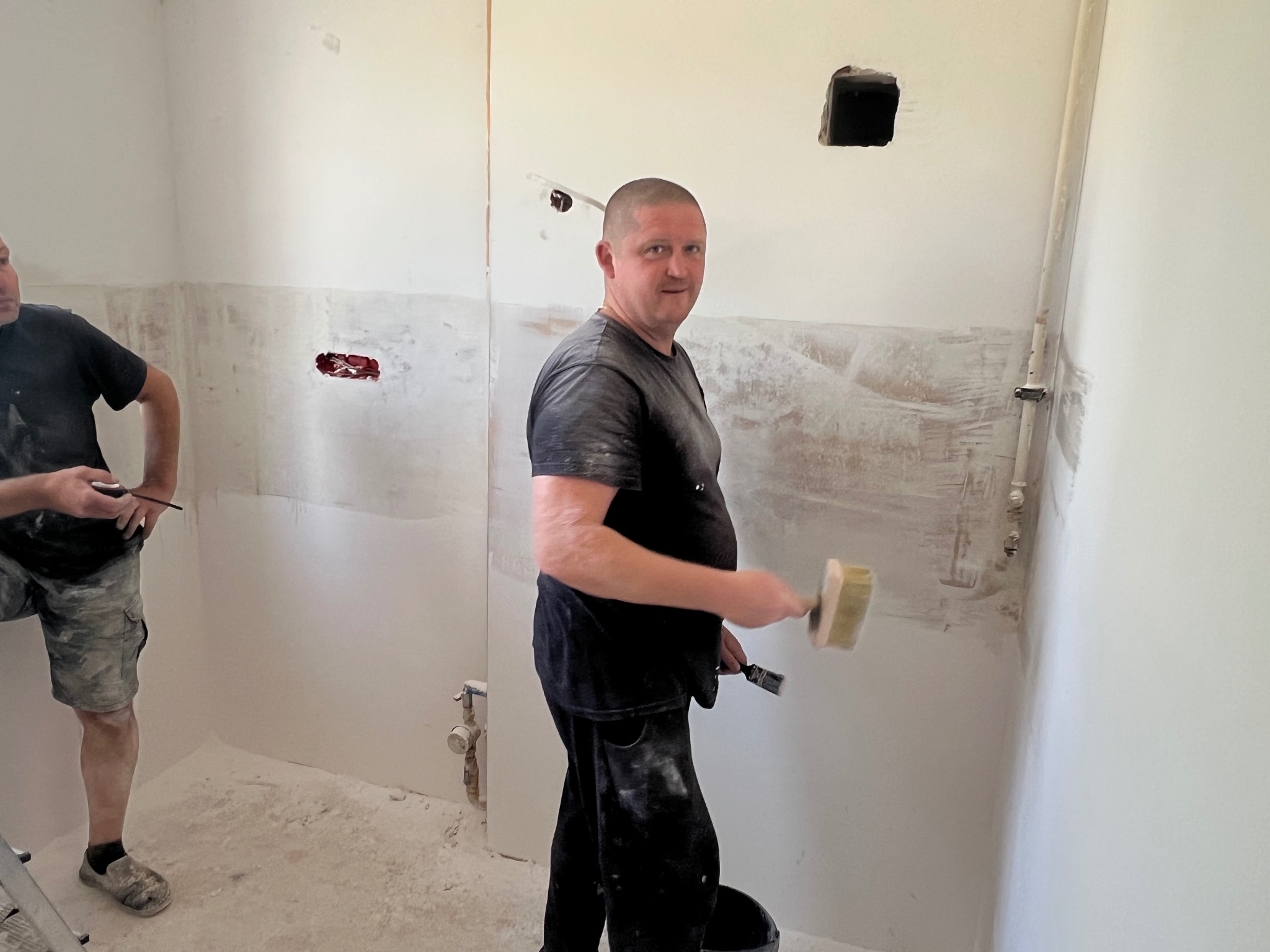 Mężczyzna z pędzlem w trakcie nakładania gładzi na ścianę w remontowanym pomieszczeniu. Widoczne ślady po instalacjach i przygotowania do malowania.