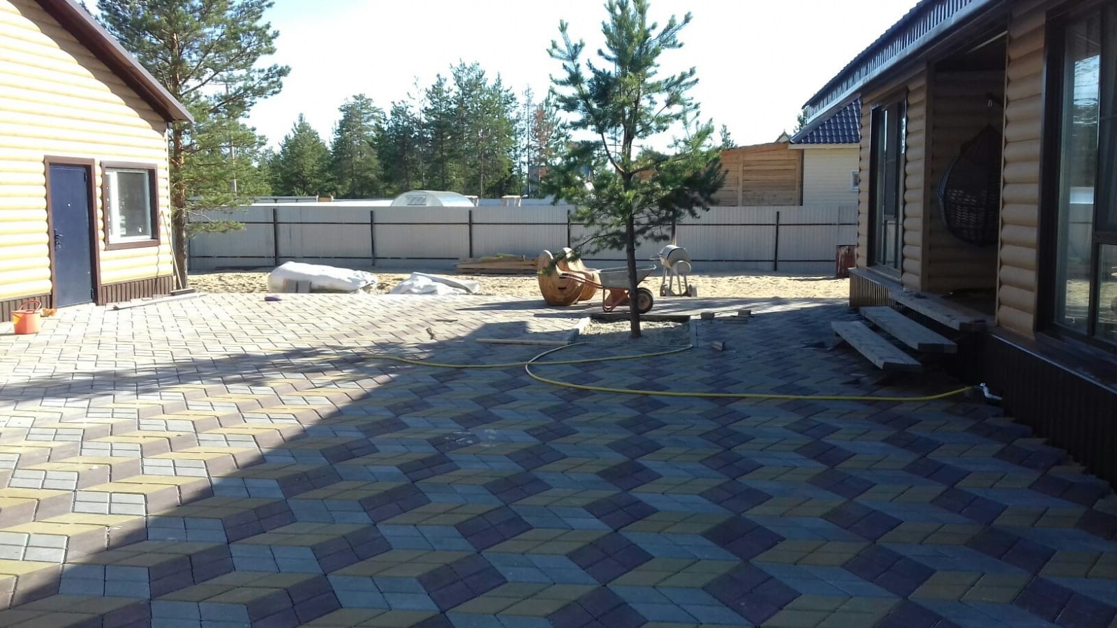 Ułożona kostka brukowa w geometryczny wzór 3D na podwórku z dwoma drewnianymi budynkami, betoniarką i taczką w tle, ogrodzenie z blachy trapezowej w oddali.